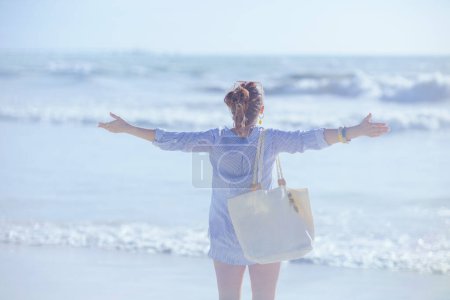 Foto de Visto desde atrás mujer con bolsa de paja blanca regocijándose en la playa. - Imagen libre de derechos