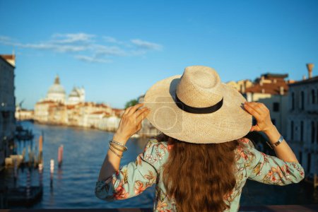 Téléchargez les photos : Seen from behind trendy woman in floral dress with hat on Accademia bridge in Venice, Italy. - en image libre de droit