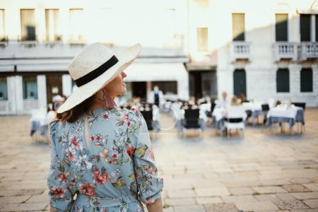 Téléchargez les photos : Seen from behind modern traveller woman in floral dress with hat sightseeing in Venice, Italy. - en image libre de droit