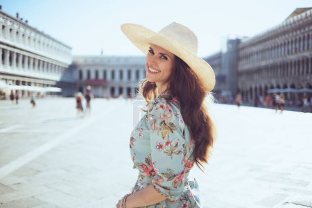 Photo for Smiling trendy middle aged traveller woman in floral dress with hat enjoying promenade at Piazza San Marco in Venice, Italy. - Royalty Free Image