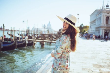 Foto de Sonriente mujer con estilo en vestido floral con sombrero teniendo recorrido a pie en terraplén en Venecia, Italia. - Imagen libre de derechos