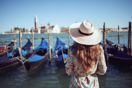 Seen from behind young woman in floral dress with hat exploring attractions on embankment in Venice, Italy.