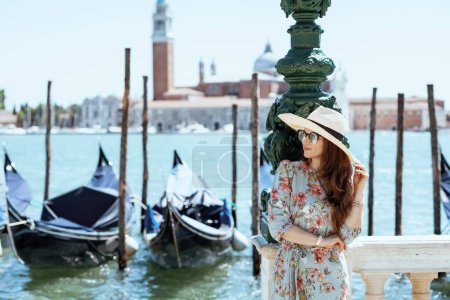 Foto de Smiling elegant solo traveller woman in floral dress with sunglasses and hat on embankment in Venice, Italy. - Imagen libre de derechos
