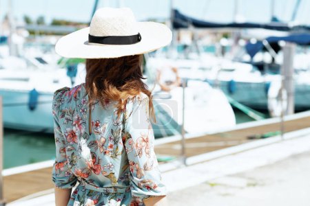 Photo for Seen from behind elegant traveller woman in floral dress with hat on the pier. - Royalty Free Image