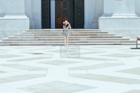 Foto de Stylish middle aged traveller woman in floral dress with hat and medical mask on San Giorgio Maggiore island. - Imagen libre de derechos