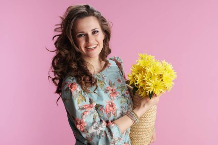 Portrait of smiling modern middle aged woman in floral dress with yellow chrysanthemums flowers and straw bag isolated on pink background.