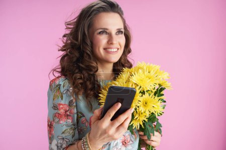 Téléchargez les photos : Smiling trendy woman in floral dress with yellow chrysanthemums flowers using smartphone applications isolated on pink. - en image libre de droit