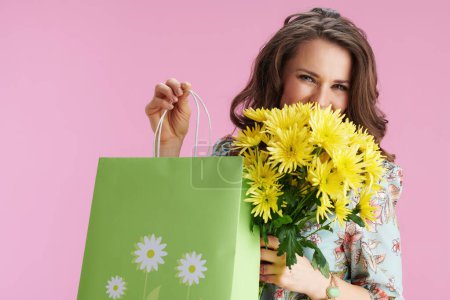 Foto de Feliz joven con el pelo largo y ondulado morena con flores de crisantemos amarillos y bolsa verde aislada sobre fondo rosa. - Imagen libre de derechos