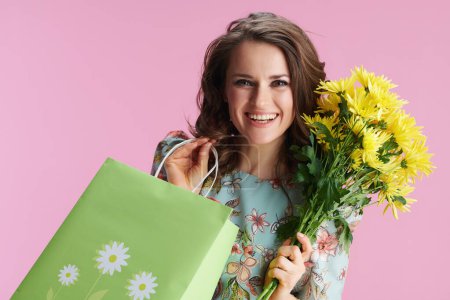 Photo for Portrait of happy stylish middle aged woman in floral dress with yellow chrysanthemums flowers and green shopping bag against pink background. - Royalty Free Image