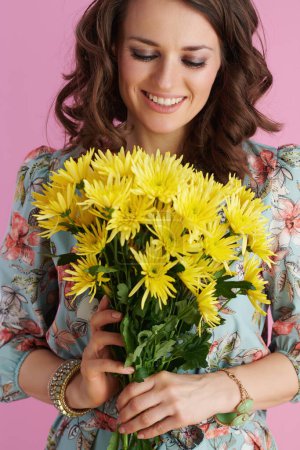 Foto de Smiling modern middle aged woman in floral dress with yellow chrysanthemums flowers against pink background. - Imagen libre de derechos