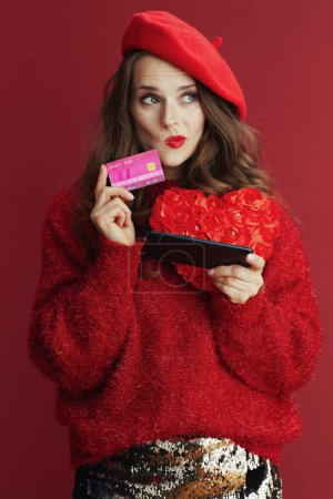 Téléchargez les photos : Happy Valentine. pensive modern woman in red sweater and beret with red heart, smartphone and credit card browsing online shop. - en image libre de droit
