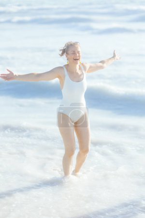 Téléchargez les photos : Full length portrait of happy elegant 40 years old woman in white swimsuit at the beach having fun time. - en image libre de droit