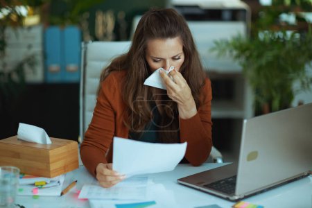 Photo for Trendy small business owner woman with laptop and napkin blowing nose and working with documents in the modern green office. - Royalty Free Image