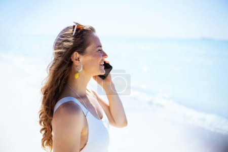 Foto de Happy stylish middle aged woman at the beach in white swimwear talking on a smartphone. - Imagen libre de derechos
