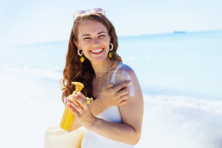 Foto de Retrato de mujer de 40 años sonriente y elegante en la playa en ropa de playa blanca aplicando spf. - Imagen libre de derechos