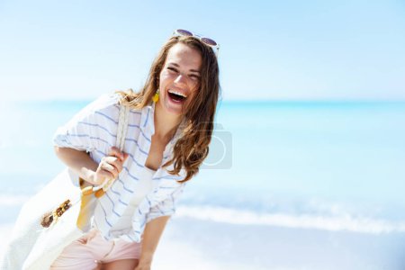 Photo for Happy stylish woman with white straw bag in white striped shirt and shorts having fun time at the beach. - Royalty Free Image