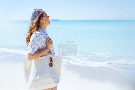 Photo for Happy stylish middle aged woman with white straw bag in white striped shirt and shorts at the beach. - Royalty Free Image
