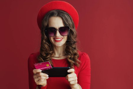 Foto de Happy Valentine. happy modern 40 years old woman with long wavy hair in red dress and beret isolated on red background with credit card and smartphone making online shopping on e-commerce site. - Imagen libre de derechos