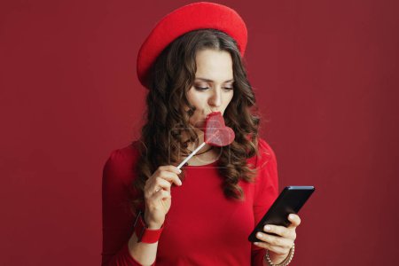Téléchargez les photos : Happy Valentine. modern woman with long wavy hair in red dress and beret on red background with heart shaped candy on stick using smartphone app. - en image libre de droit