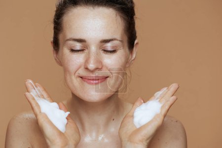 Téléchargez les photos : Happy 40 years old woman with foaming face wash washing face against beige background. - en image libre de droit