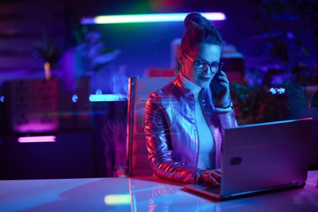 Téléchargez les photos : Neon metaverse futuristic concept. stylish business woman in glasses using laptop and using a smartphone in modern office. - en image libre de droit