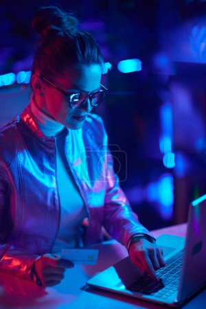 Foto de Neon metaverse futuristic concept. modern woman in glasses with credit card using laptop in modern office. - Imagen libre de derechos