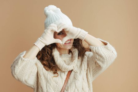 Foto de Hello winter. smiling modern 40 years old woman in beige sweater, mittens and hat showing heart shaped hands isolated on beige. - Imagen libre de derechos