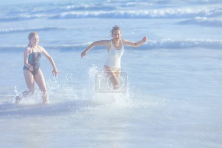 Foto de Full length portrait of happy modern mother and teenage daughter at the beach in swimwear running. - Imagen libre de derechos