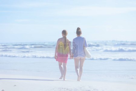 Téléchargez les photos : Vu de derrière la mère élégante et la fille adolescente à la plage marchant. - en image libre de droit