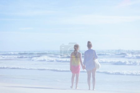 Téléchargez les photos : Vu de derrière la mère élégante et la fille adolescente à la plage regardant au loin. - en image libre de droit