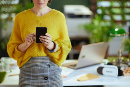 Téléchargez les photos : Gros plan sur propriétaire de petite entreprise femme en pull jaune dans le bureau vert en utilisant smartphone. - en image libre de droit