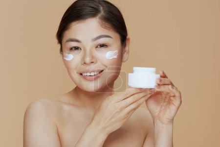 Téléchargez les photos : Portrait of modern asian woman with facial cream jar and facial cream on face on beige background. - en image libre de droit