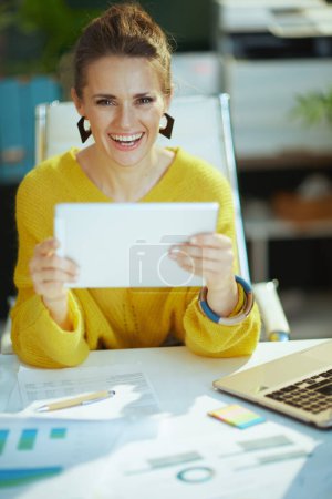 Foto de Happy trendy middle aged small business owner woman in yellow sweater with tablet PC in the modern green office. - Imagen libre de derechos
