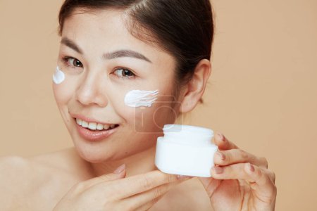 Téléchargez les photos : Portrait of modern asian female with facial cream jar and facial cream on face on beige background. - en image libre de droit