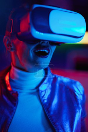Téléchargez les photos : Neon metaverse futuristic concept. Portrait of surprised stylish middle aged woman in vr headset on dark background. - en image libre de droit