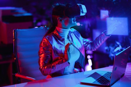 Téléchargez les photos : Neon metaverse concept futuriste. curieux élégant 40 ans femme en réalité virtuelle lunettes en utilisant la technologie de réalité virtuelle dans le bureau moderne. - en image libre de droit