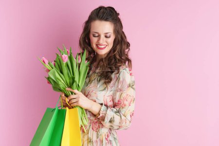 Téléchargez les photos : Femme élégante et souriante en robe florale avec bouquet de tulipes et sacs à provisions isolés sur rose. - en image libre de droit