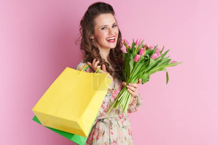 Photo for Smiling elegant woman in floral dress with tulips bouquet and shopping bags isolated on pink background. - Royalty Free Image