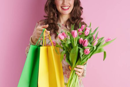 Téléchargez les photos : Gros plan sur femme souriante en robe florale avec bouquet de tulipes et sacs à provisions isolés sur fond rose. - en image libre de droit