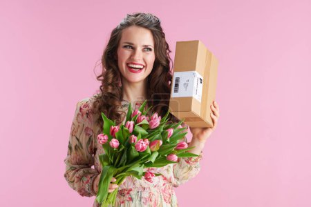 Photo for Smiling stylish middle aged woman with long wavy brunette hair with tulips bouquet and parcel isolated on pink background. - Royalty Free Image