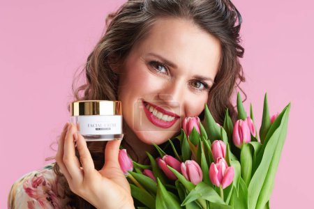 Téléchargez les photos : Portrait de femme moderne souriante en robe florale avec bouquet de tulipes et crème faciale isolée sur fond rose. - en image libre de droit