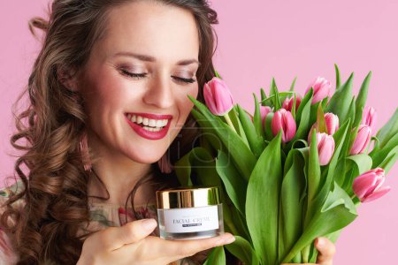 Téléchargez les photos : Femme élégante et souriante en robe florale avec bouquet de tulipes et crème faciale isolée sur fond rose. - en image libre de droit