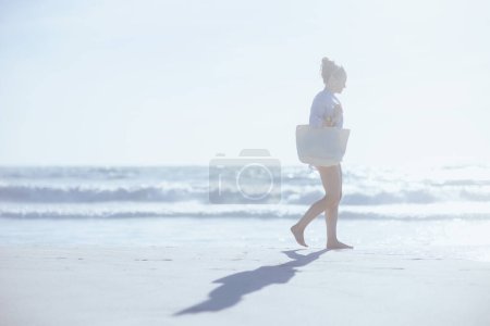 Téléchargez les photos : Portrait pleine longueur de heureuse femme d'âge moyen moderne avec sac de paille blanche marchant à la plage. - en image libre de droit