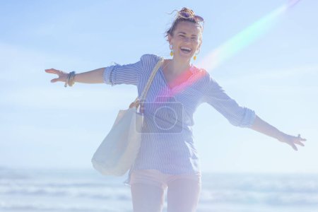 Téléchargez les photos : Femme souriante élégante de 40 ans avec sac de paille blanche se réjouissant à la plage. - en image libre de droit