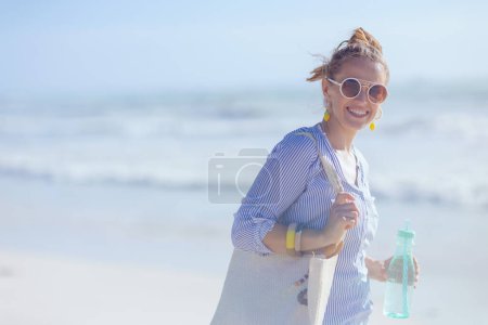 Téléchargez les photos : Heureuse femme élégante avec sac de paille blanche et boisson à la plage. - en image libre de droit