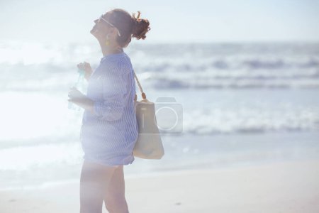 Photo for Smiling elegant 40 years old woman with white straw bag and drink at the beach. - Royalty Free Image