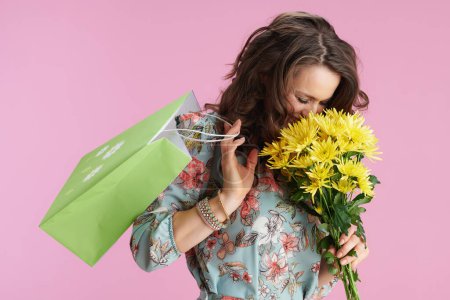 sonriente mujer de moda con el pelo largo y ondulado morena con flores de crisantemos amarillos y bolsa de compras verde sobre fondo rosa.