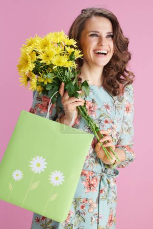Photo for Smiling elegant middle aged woman in floral dress with yellow chrysanthemums flowers and green shopping bag isolated on pink background. - Royalty Free Image