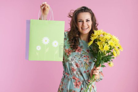 Téléchargez les photos : Happy elegant woman with long wavy brunette hair with yellow chrysanthemums flowers and green shopping bag isolated on pink background. - en image libre de droit