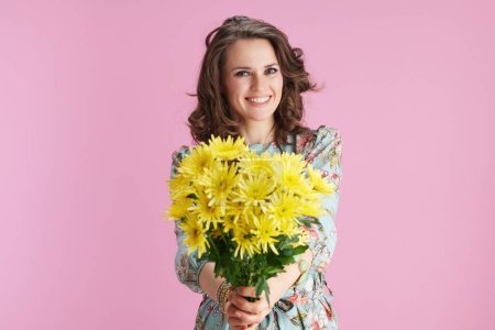 Photo for Portrait of smiling young female in floral dress giving yellow chrysanthemums flowers against pink background. - Royalty Free Image